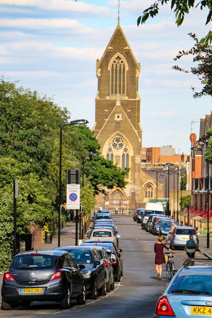 A residential street in Dollis Hill featuring a row of red-brick Victorian-style terraced houses with white window frames and pitched roofs. The street is lined with parked cars on both sides, includi