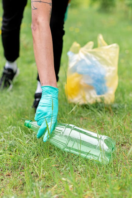 A person wearing a blue rubber glove is picking up a green plastic bottle from a grassy area during a cleanup or gardening activity. In the background, there is a yellow plastic bag filled with other waste or recyclables, placed on the grass, indicating an environmentally conscious effort. The scene suggests outdoor waste collection, possibly as part of a property clearance or community clean-up, which aligns with moving or decluttering tasks that Man with Van Dollis Hill offers in home relocations or furniture transport. The image captures the outdoor environment with natural lighting, focusing on the act of environmental responsibility and preparing for proper waste disposal, relevant to the broader themes of packing, moving logistics, and responsible waste management related to house removals.