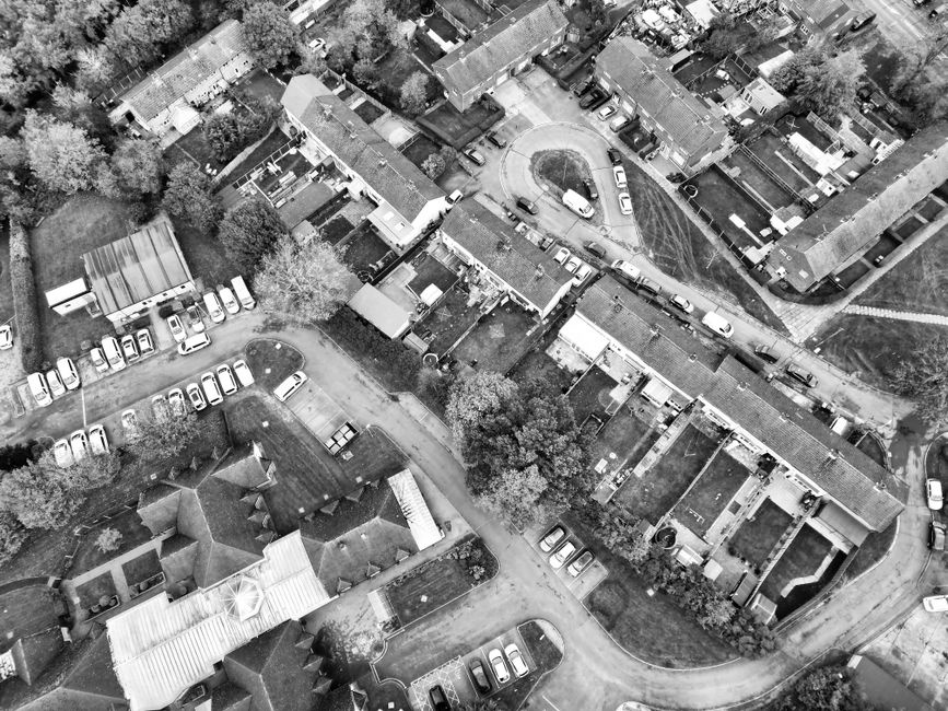 An aerial black-and-white photograph of a residential area showing multiple terraced houses with pitched roofs, surrounded by trees and small garden yards. In front of the houses, a narrow street is visible with parked cars along the pavement and a small roundabout at a junction. Near the bottom left, a house with a conservatory and a driveway is present, with several cars parked nearby. In the upper part of the image, a group of large cardboard boxes, wrapped furniture, and secured household items are being loaded onto a moving van parked on the street, with visible straps and blankets for protection. ManwithVandollishill.co.uk, providing professional removals in the area, is involved in the moving process, which includes careful packing and transport of household contents during a house relocation or furniture transport. The scene emphasizes the logistics of professional moving services, including careful handling of furniture and boxes in an outdoor environment at daylight.