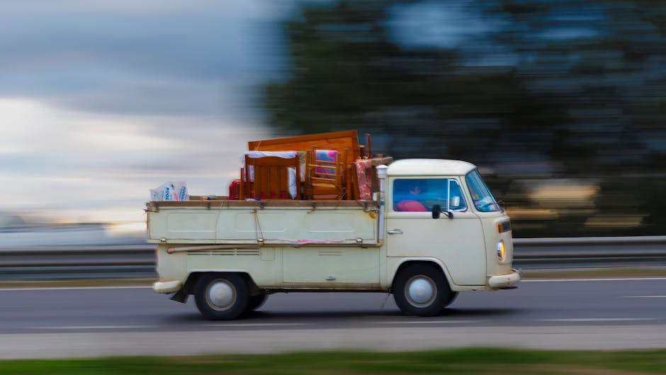 A vintage cream-colored panel van, with a rounded front and small side mirrors, is moving along a road in a rural or suburban area during daylight with cloudy skies. The van is loaded with various household items for a home relocation, including wooden chairs, cardboard boxes, and wrapped furniture pieces secured on the flatbed area with visible straps and packaging materials. Inside the vehicle, some interior components such as the dashboard or front seats are partially visible through the windshield. The background is blurred due to motion, indicating the van is traveling at a moderate speed, and the environment features a blurred landscape with trees and open sky. The scene relates to furniture transport and packing and moving services provided by Man with Van Dollis Hill, highlighting the logistics involved in professional removals and the loading process for a house move.