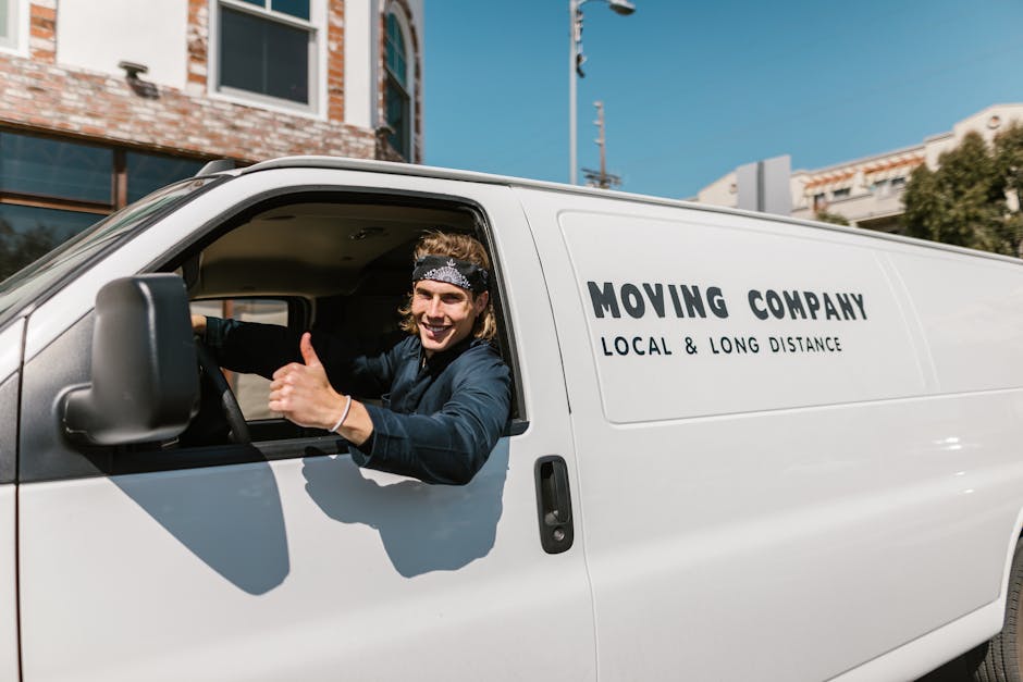 A young man with shoulder-length hair, wearing a patterned headband and dark clothing, is sitting in the driver's seat of a white moving van parked outside a residential building. He is smiling and gi