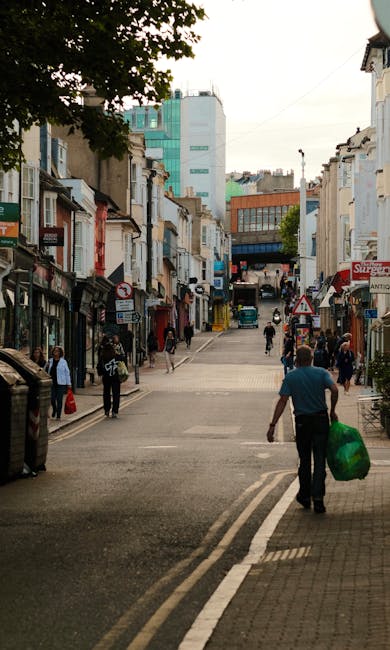 A busy street scene in Dollis Hill showing a variety of low-rise retail and residential buildings lining both sides of the pedestrianized area. The street is partially filled with people walking, some carrying shopping bags or backpacks. In the foreground, a man wearing a blue shirt and black trousers is walking away from the camera, holding a large green shopping bag. The street surface features white road markings and tactile paving at the crosswalk. Tall modern office buildings can be seen further in the background, along with a blue sky and some trees. This setting reflects an active neighbourhood suitable for local house removals or furniture transport, with no visible moving equipment, but the environment suggests a typical area where a professional removal service like Man with Van Dollis Hill might operate during home relocation activities.