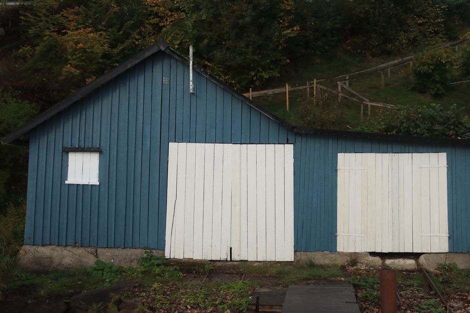Photograph of a blue wooden building with vertical paneling, featuring two white wooden shutters on the right side and a small white boarded-up window on the left. The building is situated outdoors, with a concrete foundation visible at the base and a slight incline leading up to an area of greenery and trees in the background. A metal pole extends from the roof, possibly for a weather sensor or antenna. The ground in front includes a narrow pathway, and the setting suggests a rural or semi-rural environment. The image depicts a scene relevant to house or shed clearance, with the structure potentially involved in home relocation or storage, aligned with moving services offered by Man with Van Dollis Hill.