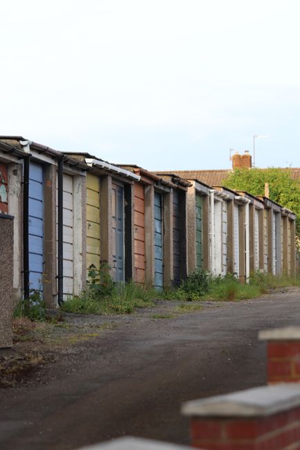 A row of small, individual storage sheds or garages aligned along a gravel pathway in Dollis Hill, with each door painted in different pastel colours including blue, pink, white, and green. The sheds have corrugated metal roofs, some with visible rust spots, and are constructed from wood and concrete panels. In the foreground, there are some cardboard boxes and packing materials, suggesting a packing or clearing process related to house removals or property clearance services. The area appears to be outdoors during the daytime with natural lighting, and there is some overgrown grass and weeds around the bases of the sheds. This setting aligns with the activities of loading, packing, or clearing during a house or garage clearance. 'Man with Van Dollis Hill' provides professional removals and packing services, including clearing of sheds and garages, as seen in this environment.