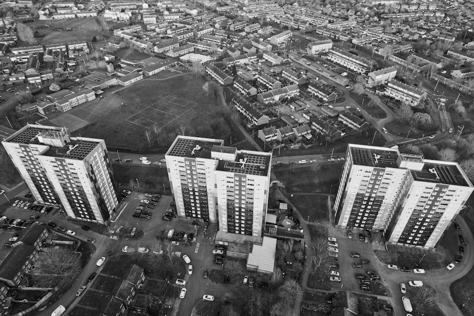 A black-and-white aerial photograph showing three tall, modern residential apartment buildings with multiple windows and flat roofs, situated in a suburban area with nearby parking lots filled with cars and surrounded by lower-rise houses, green spaces, and tree-lined streets. The foreground features a parking area adjacent to the apartment blocks, with several vehicles parked along the perimeter. In the middle ground, there are pathways, small gardens, and landscaped areas, while the background reveals a mix of single-family homes, additional residential buildings, open fields, and recreational areas, all under diffused natural light. The scene captures the urban environment and facilitates understanding of the space for home relocation discussions, with visual cues relevant to furniture transport and the logistics involved in moving processes, as seen in professional removals services such as those provided by Man with Van Dollis Hill.