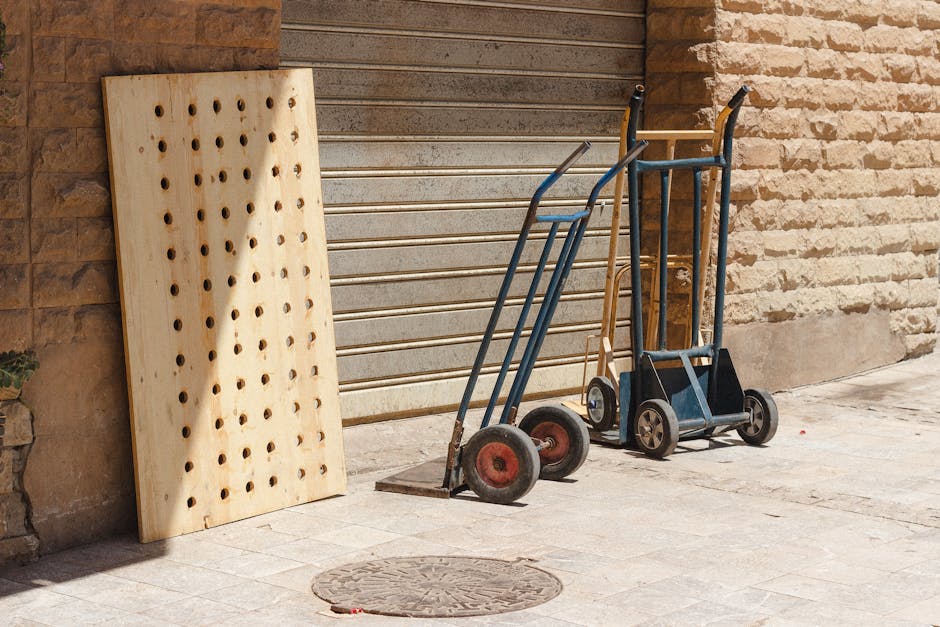 Outside a property with a brick wall and a metal gate, three hand trucks and a small trolley are positioned on the pavement near the entrance. The first hand truck has a black frame with red wheels, the second features a blue frame with black wheels, and the third has a rusted metal frame with black wheels. A wooden pegboard with multiple holes is leaning against the brick wall. The scene suggests preparation for a home relocation or furniture transport, with the equipment ready for use in loading or moving belongings, in line with the services offered by Man with Van Dollis Hill. Natural daylight illuminates the area, and the ground is paved with light-colored bricks, indicating a typical urban or residential setting suitable for house removals and packing activities.