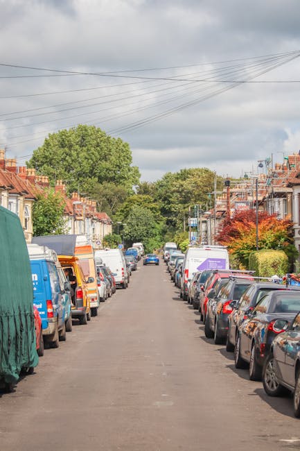 A view of a residential street in Dollis Hill with rows of parked cars on both sides, including various models and colours. The street is lined with terraced houses featuring brick facades, with some 