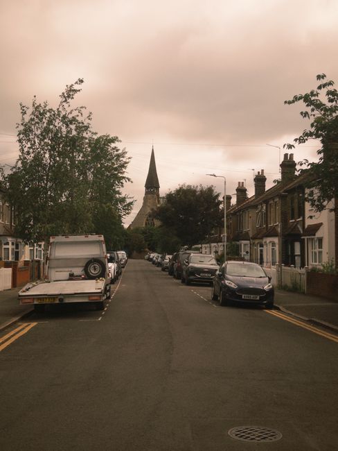 A view of a residential street in Dollis Hill with rows of parked cars on both sides, including various models and colours. The street is lined with terraced houses featuring brick facades, with some 
