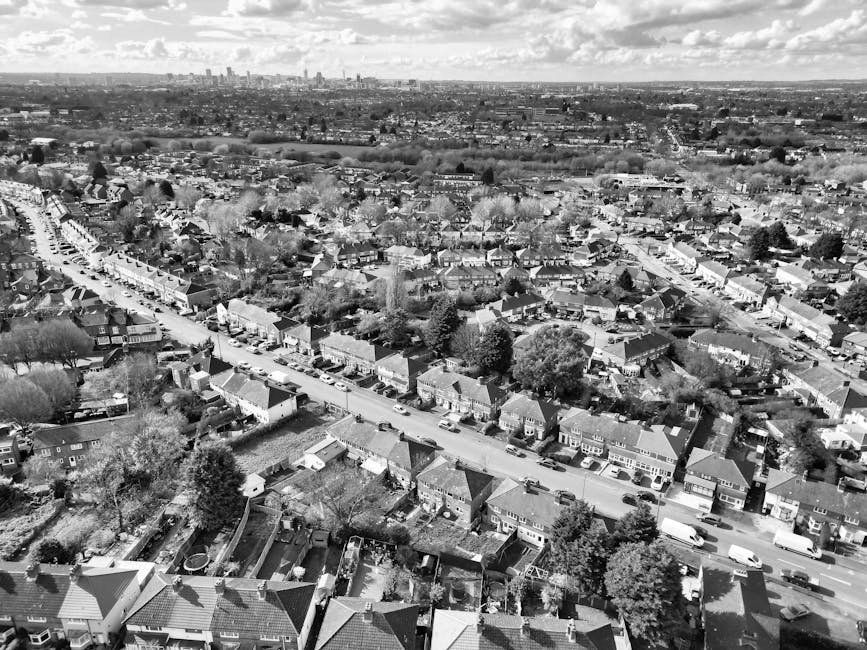 Black and white aerial photograph of a residential suburban area featuring rows of houses with pitched roofs, surrounded by trees and gardens. The street runs centrally through the image, with parked 